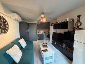 Ein kompakter Wohnraum mit Sofa, Couchtisch, Fernseher, Küchenzeile und Wanduhr.