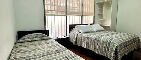 A room with two beds, a window with blinds, a wardrobe, and a ceiling light.