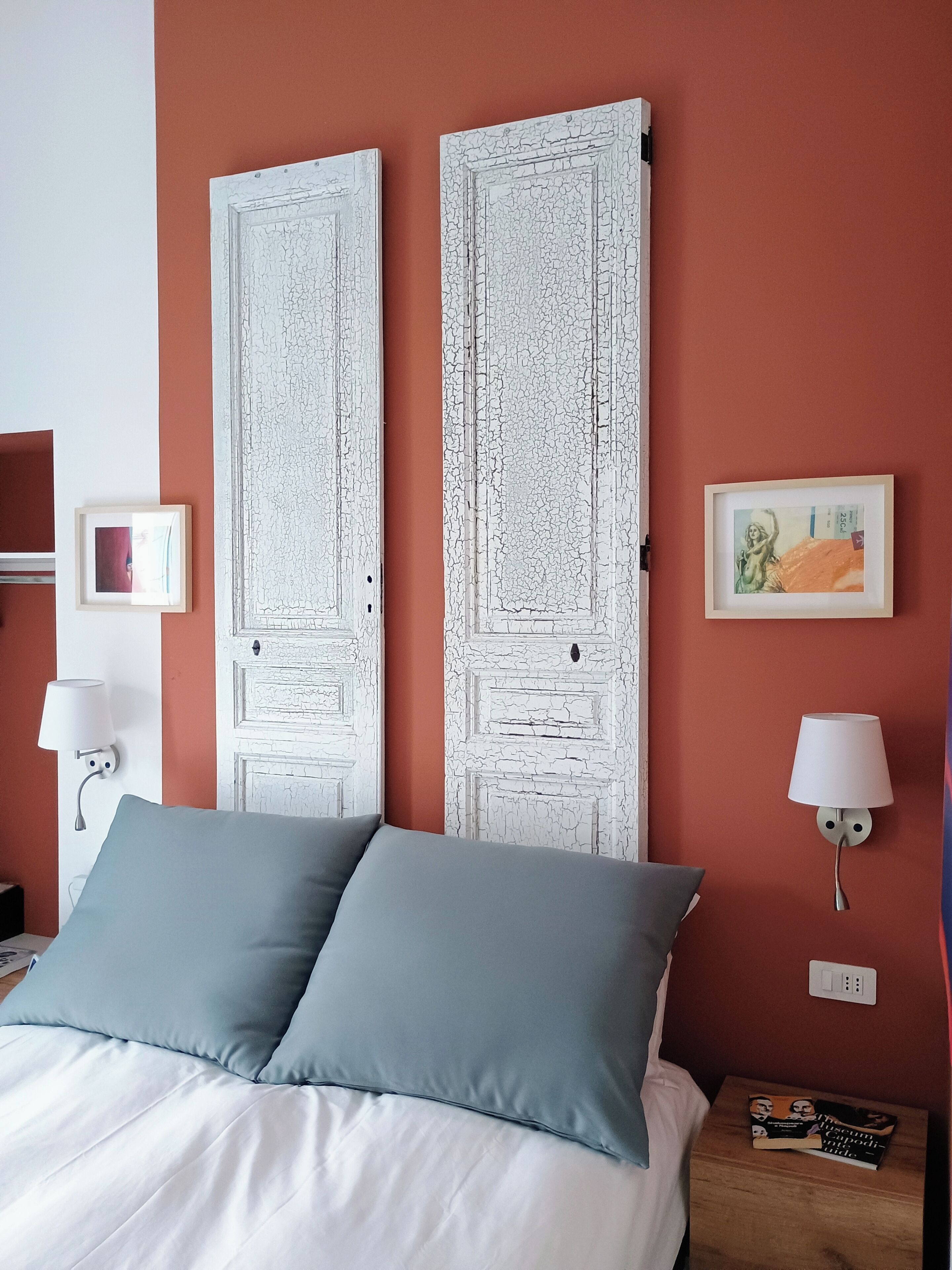 Une chambre d’hôtel comprenant un lit, deux portes, une table de chevet, deux lampes fixées au mur et deux tableaux encadrés.