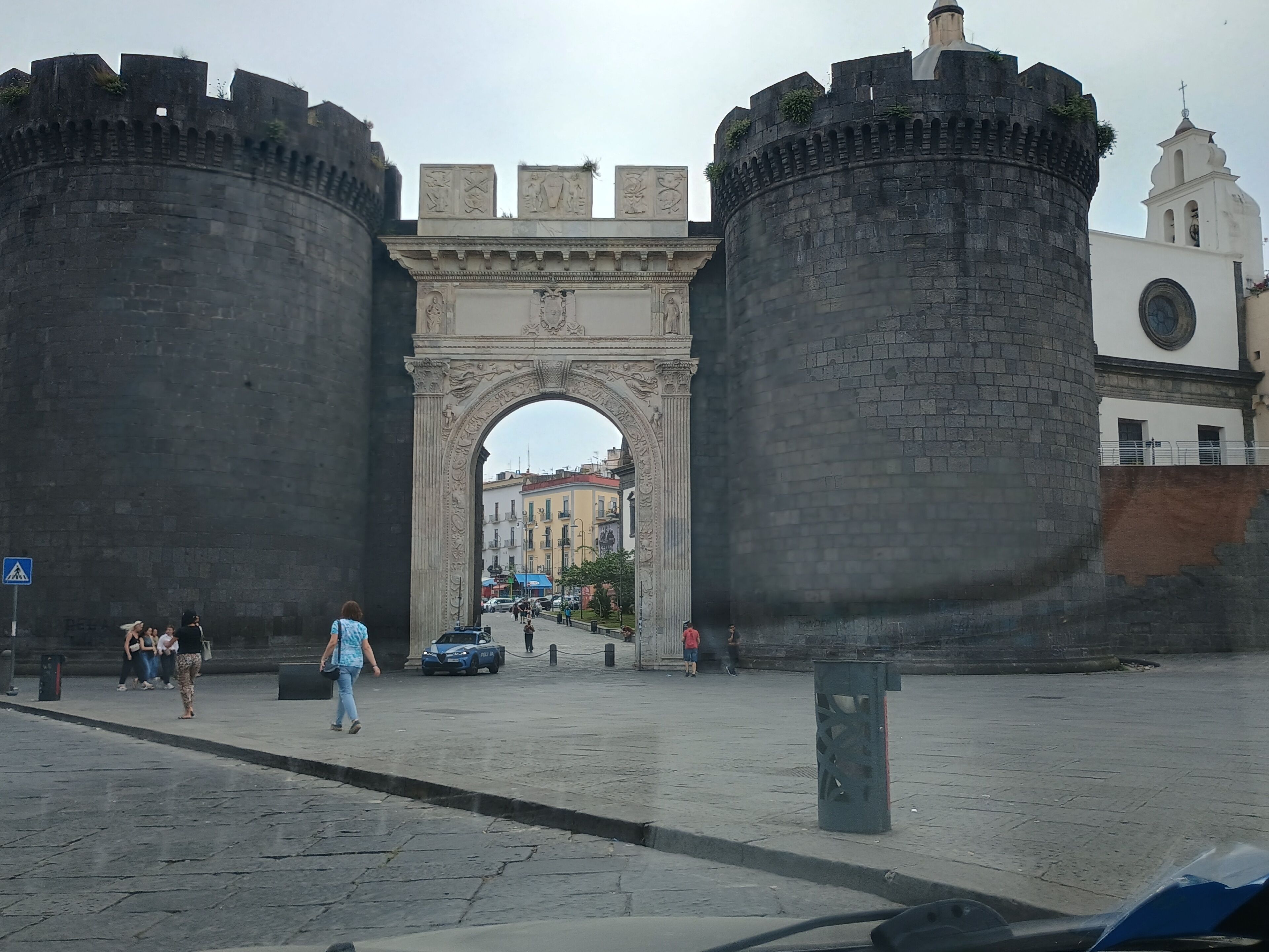 Un arc en pierre d’époque, flanqué de deux grandes tours, une place pavée et, au fond, un bâtiment blanc surmonté d’une tour horloge.