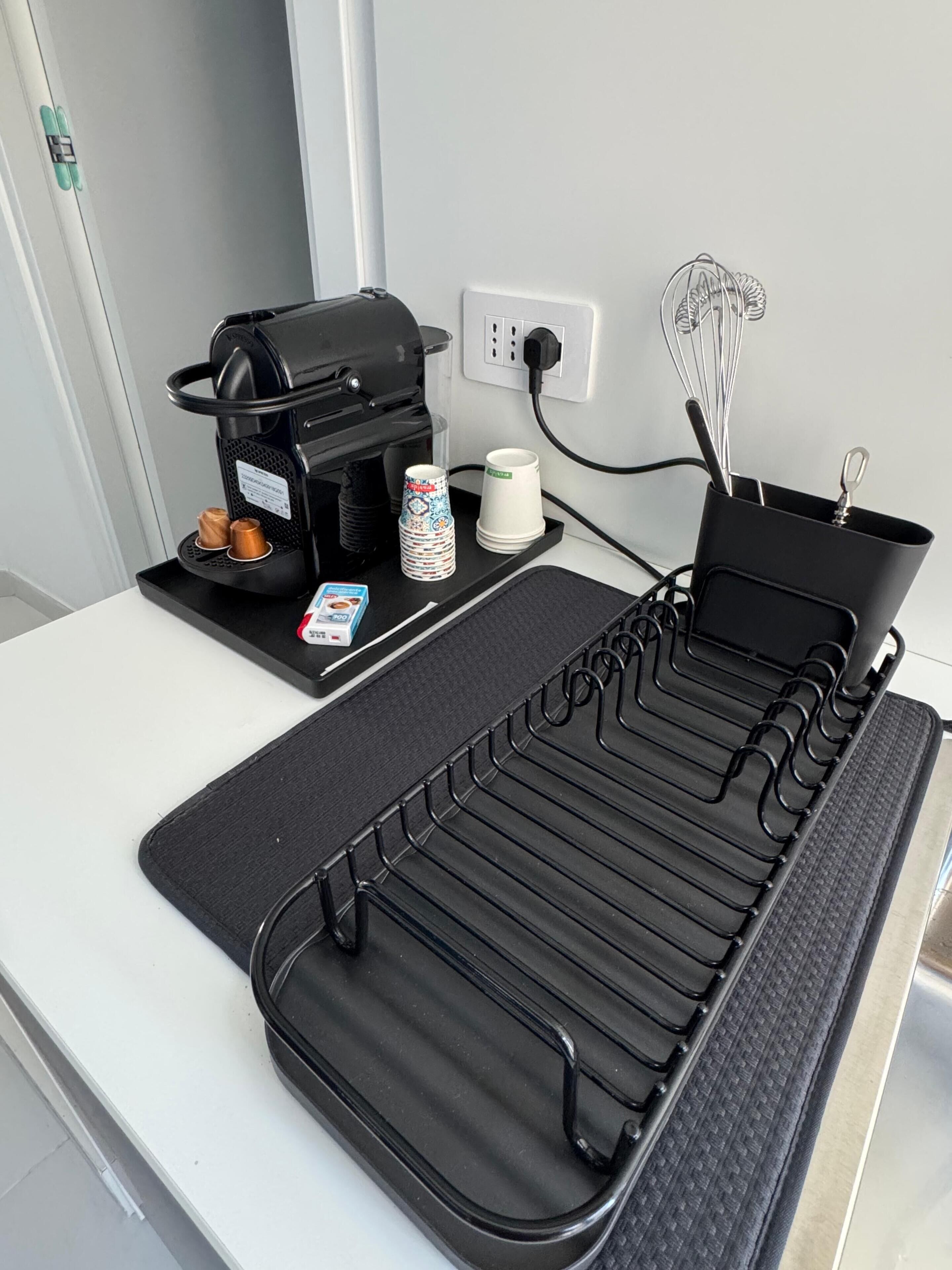 En köksbänk med en kaffebryggare, en vattenkokare, en mugg, en behållare med köksredskap och ett torkställ för brickor.