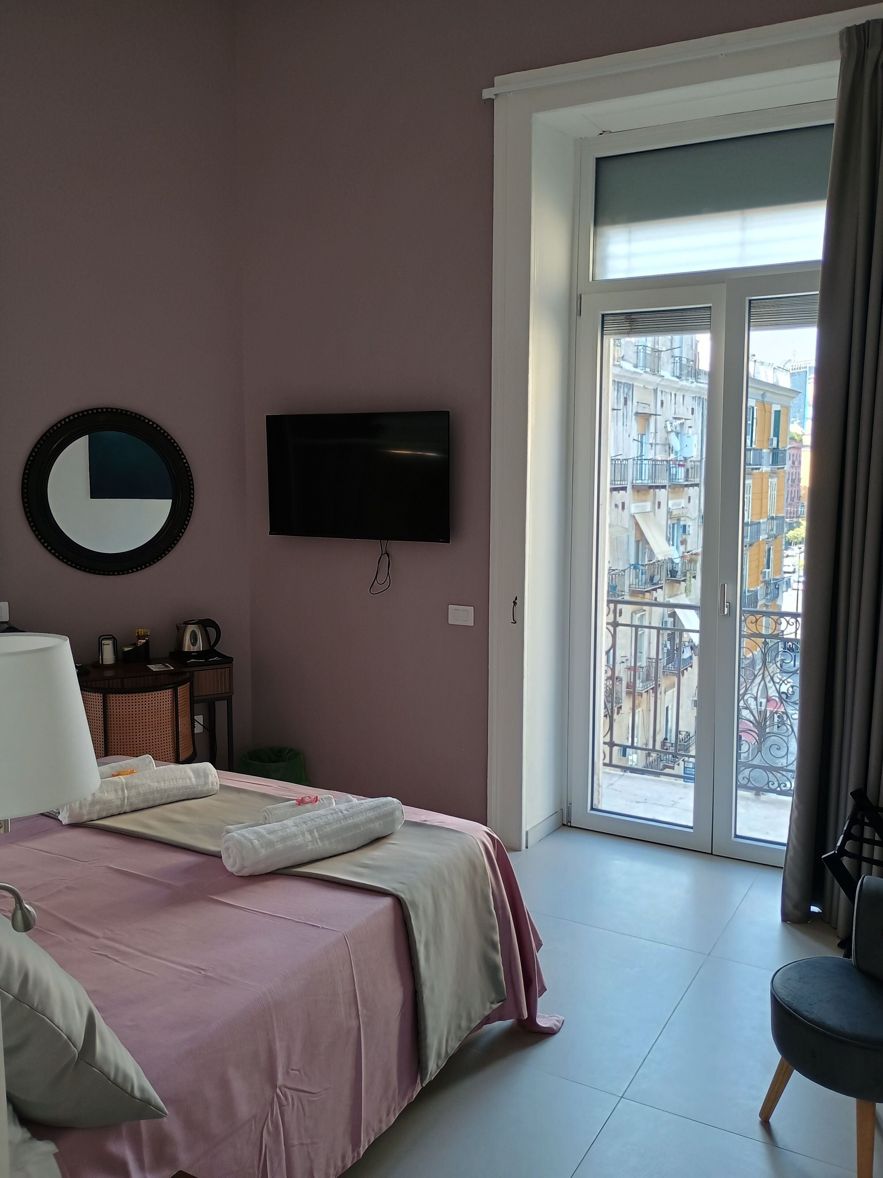 A hotel room with a bed, a TV, a window with a view of buildings, and a door.