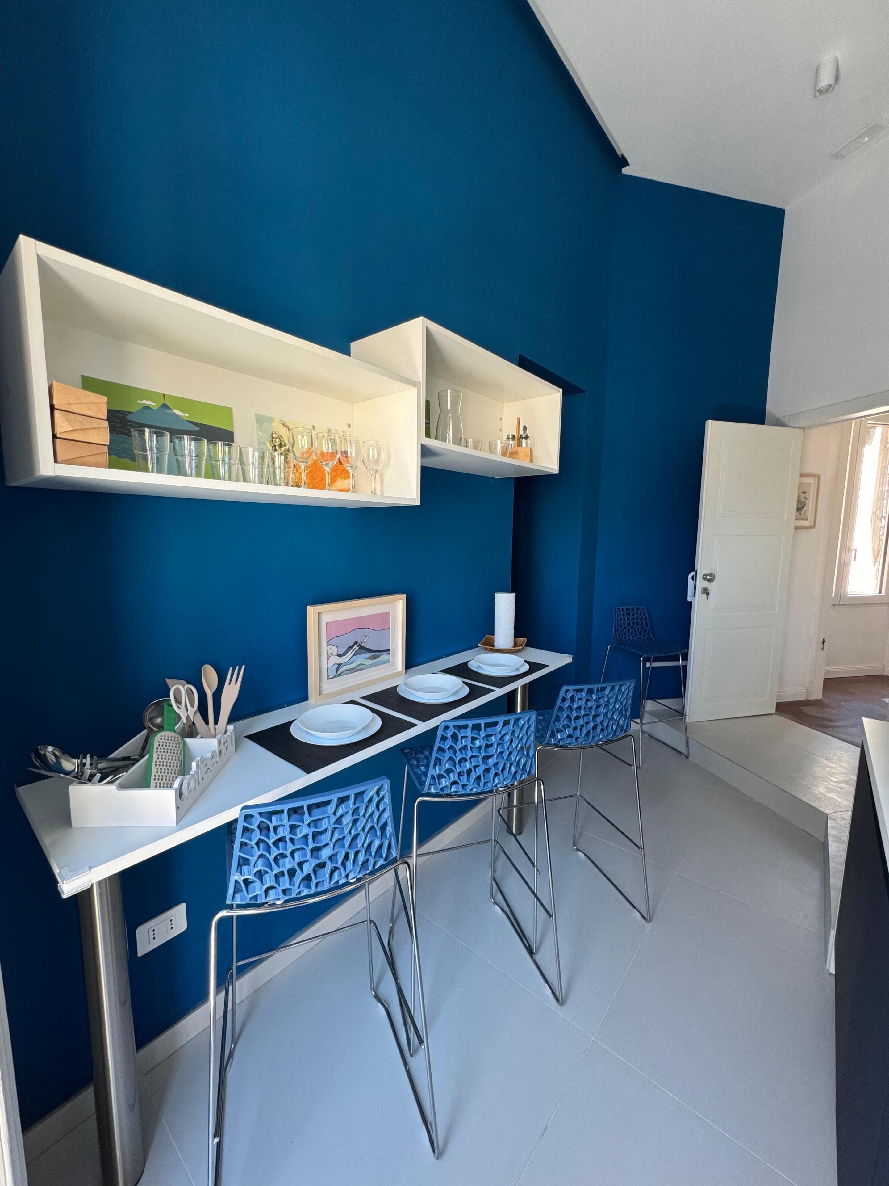 A dining area with a white table, blue chairs, and shelves above holding glasses and a painting.