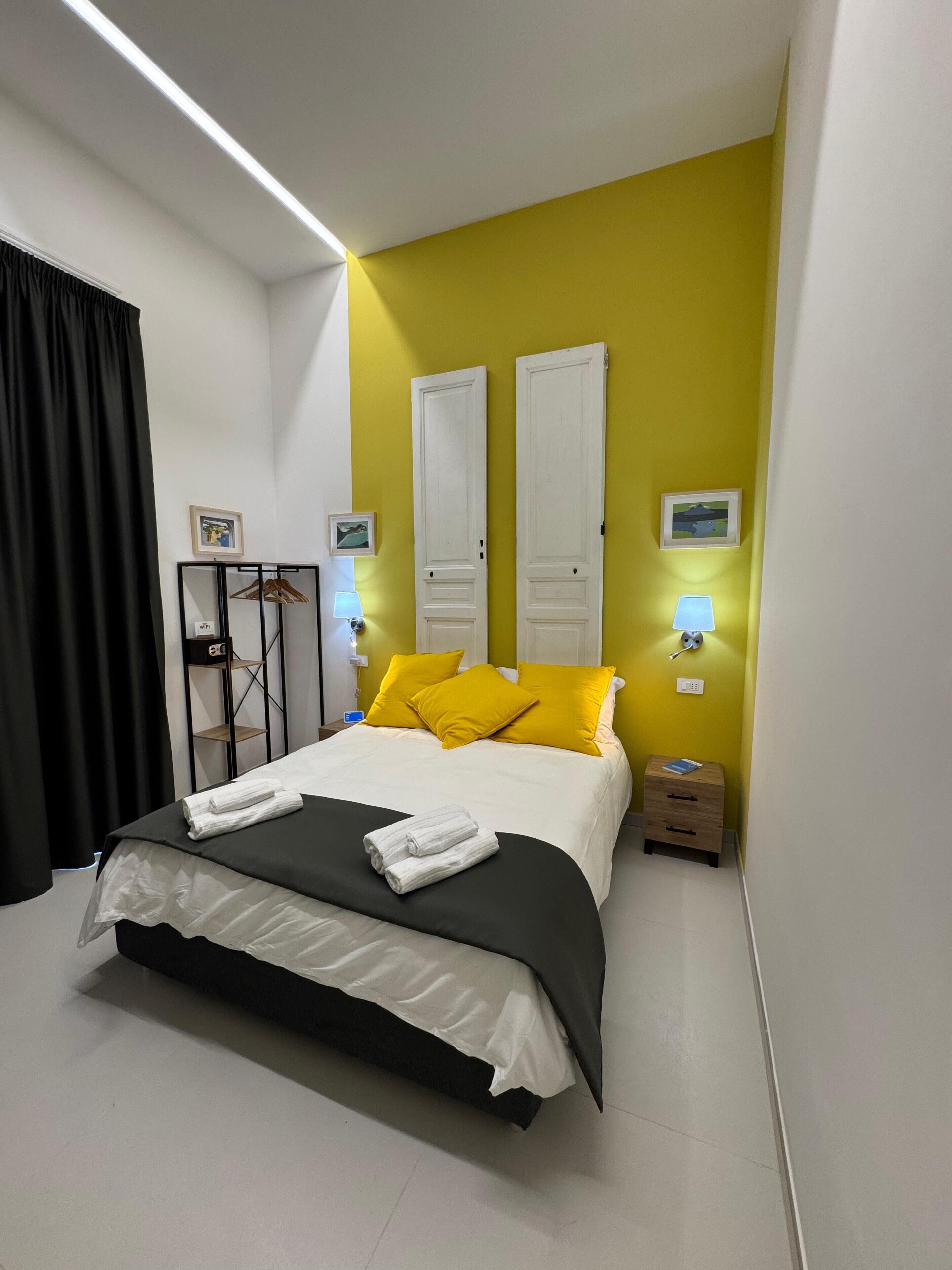 A hotel room with a bed, two bedside lamps, a nightstand, a small wooden cabinet, and a black curtain.