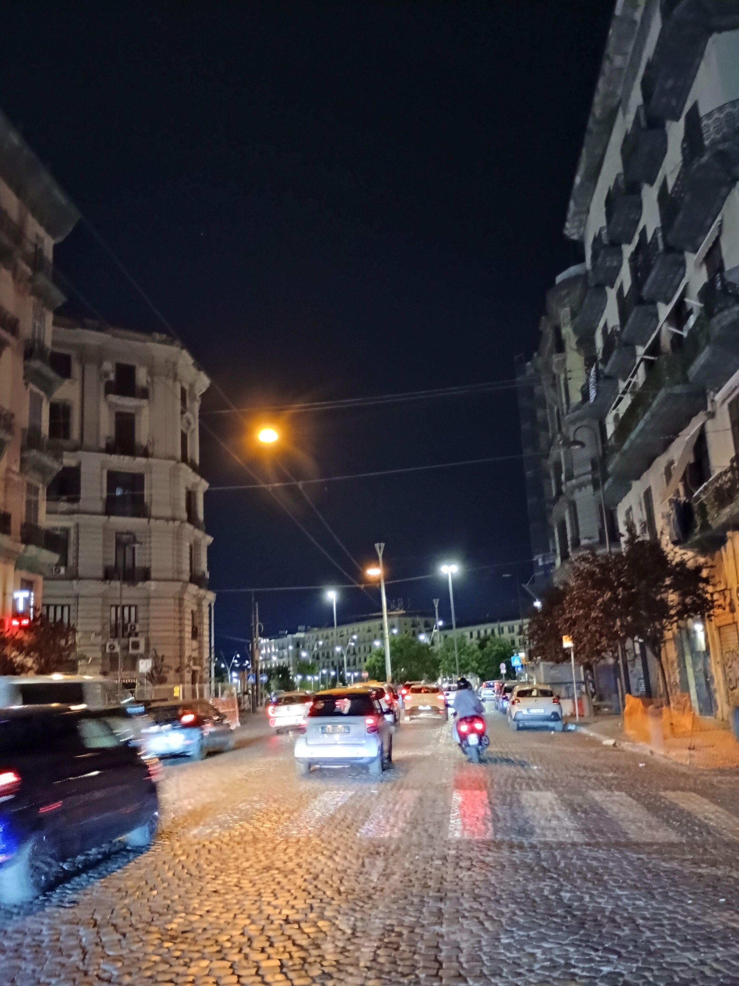 Escena nocturna de una calle con adoquines, edificios y vehículos.