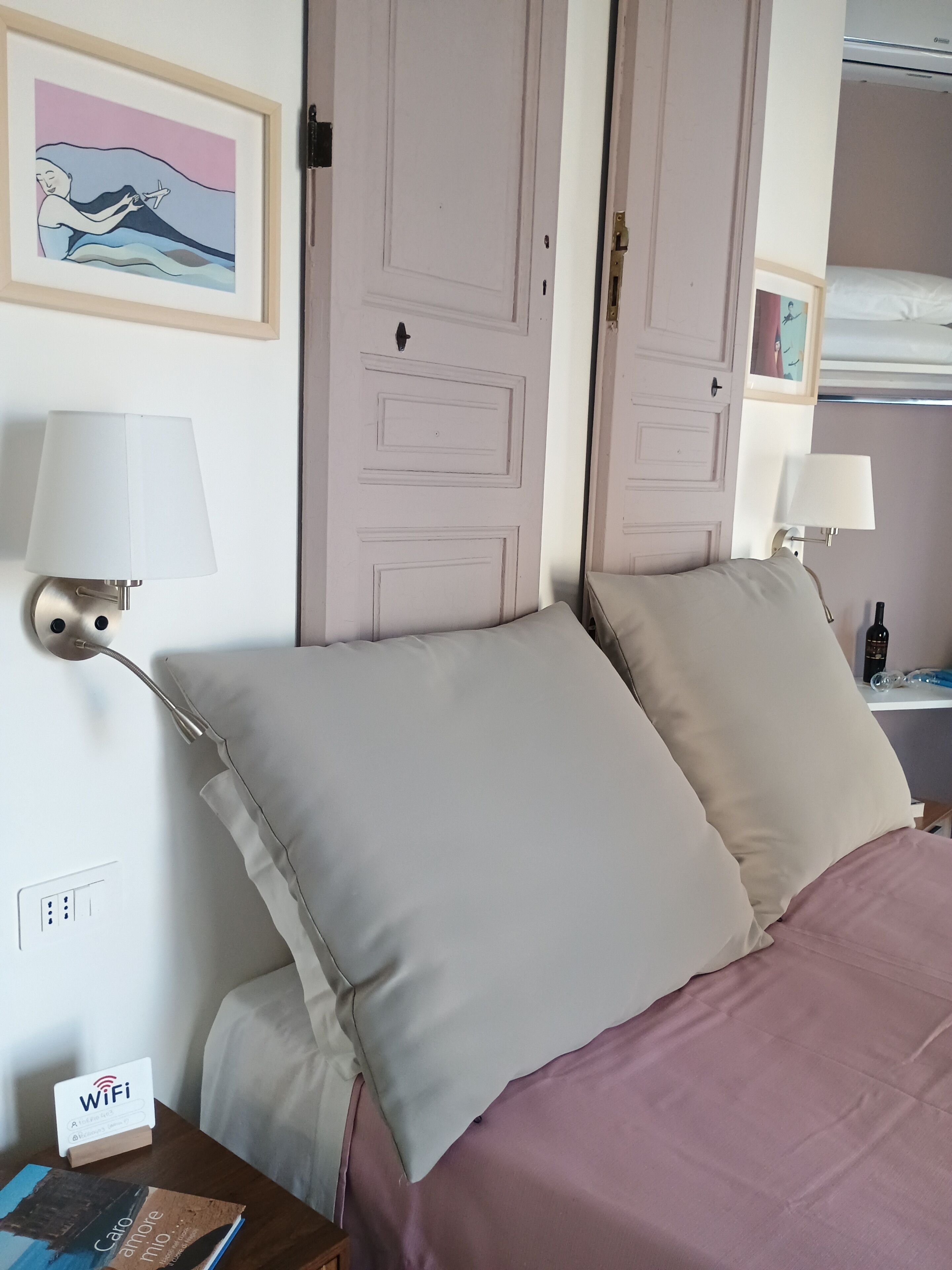 A hotel room with a bed, two bedside lamps, a framed picture on the wall, a Wi-Fi sign, and a small table with a book.
