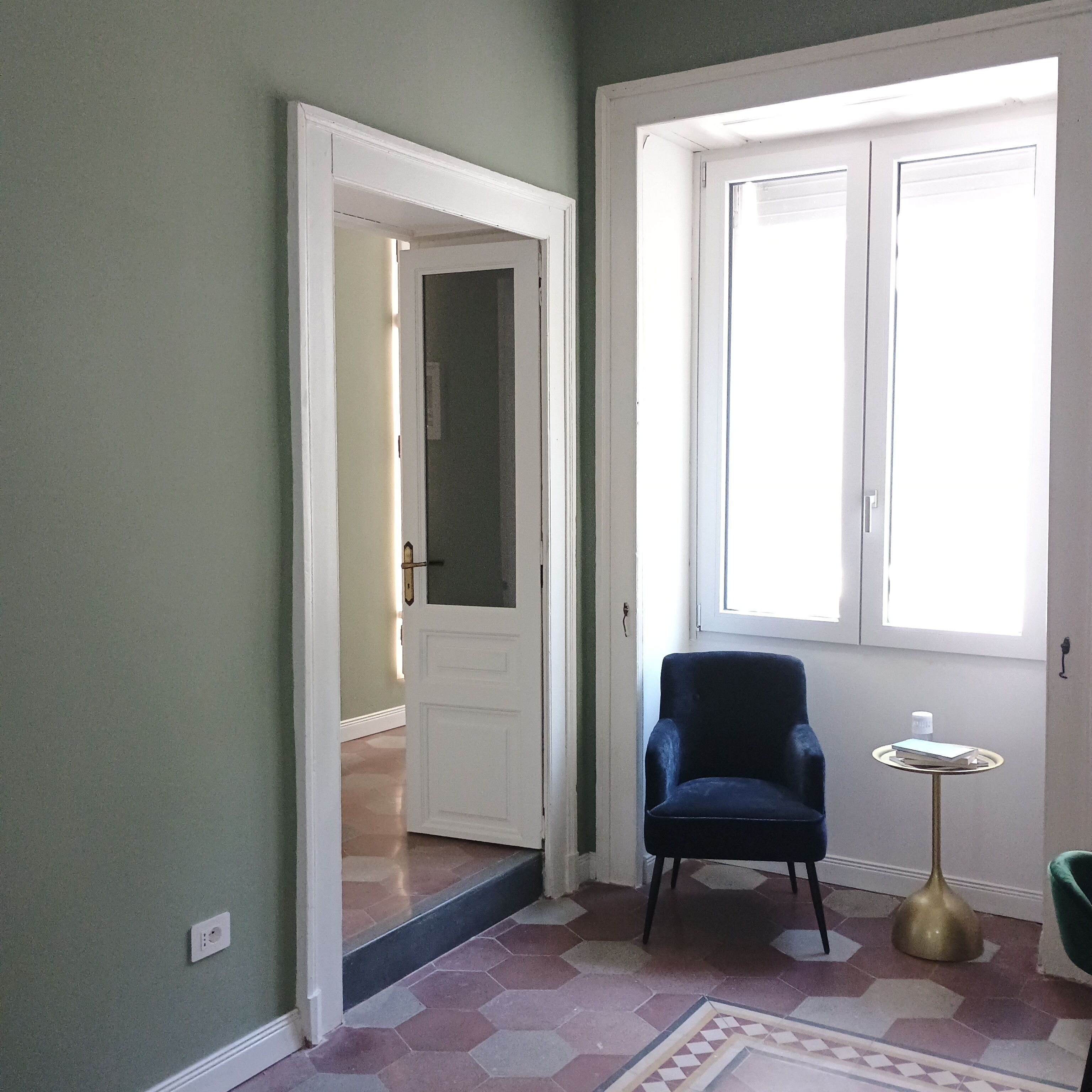A room with a door leading to another space, a window with blinds, a chair, a small table, and a patterned floor.