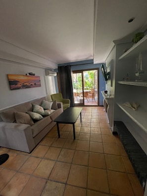 A living room with a sofa, a coffee table, and a TV on the wall. There is a sliding glass door leading to an outdoor area with a table and chairs.