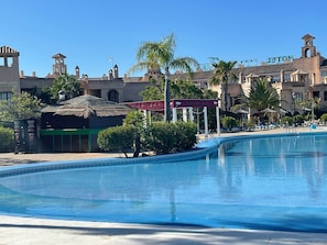 A swimming pool with a clear blue water, surrounded by palm trees and a building with a thatched roof.