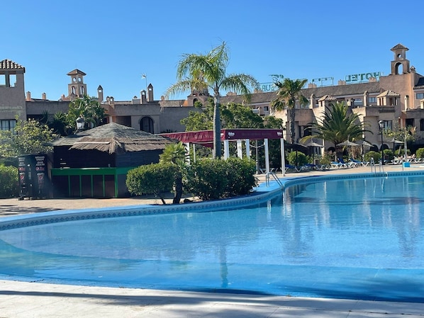 Una piscina con agua azul cristalina, rodeada de palmeras y un edificio con techo de paja.