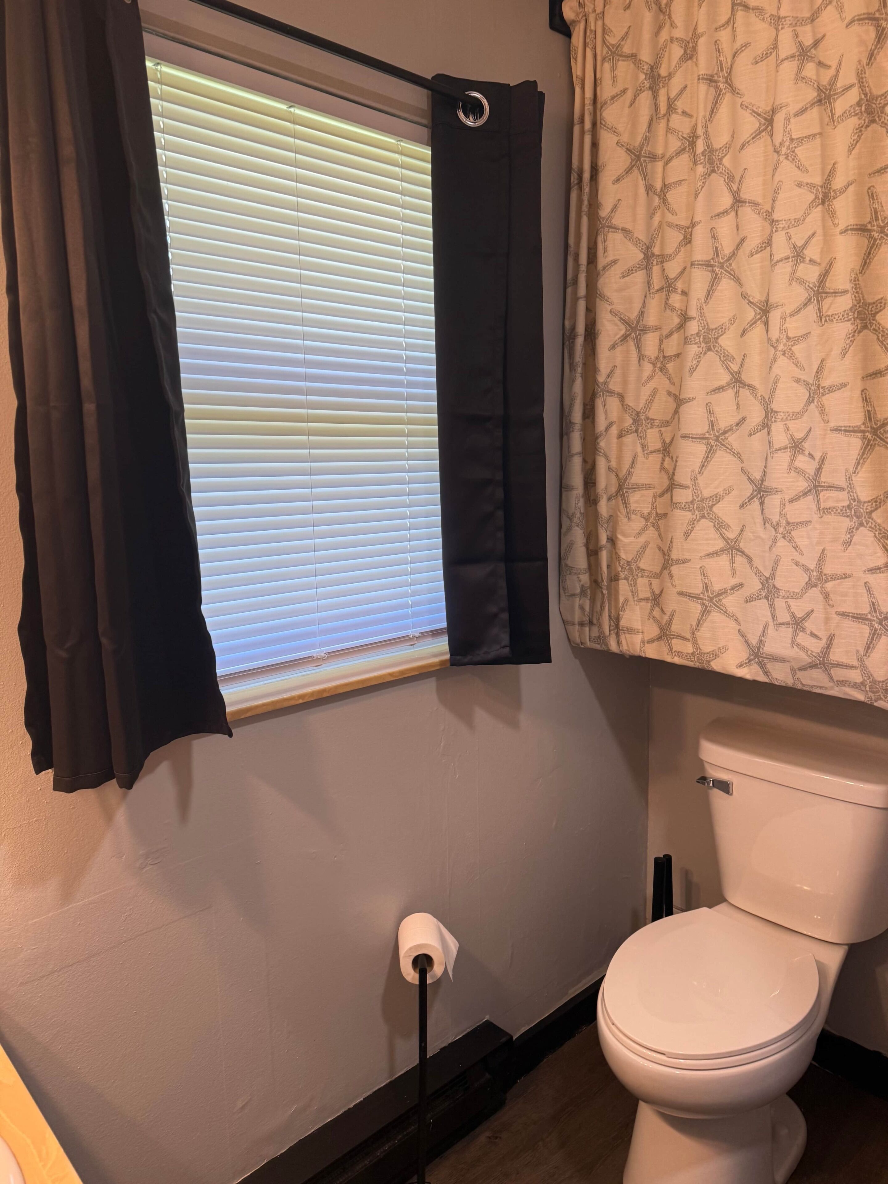 Une salle de bain comprenant des toilettes, un rouleau de papier toilette, une fenêtre avec des stores et un rideau de douche.