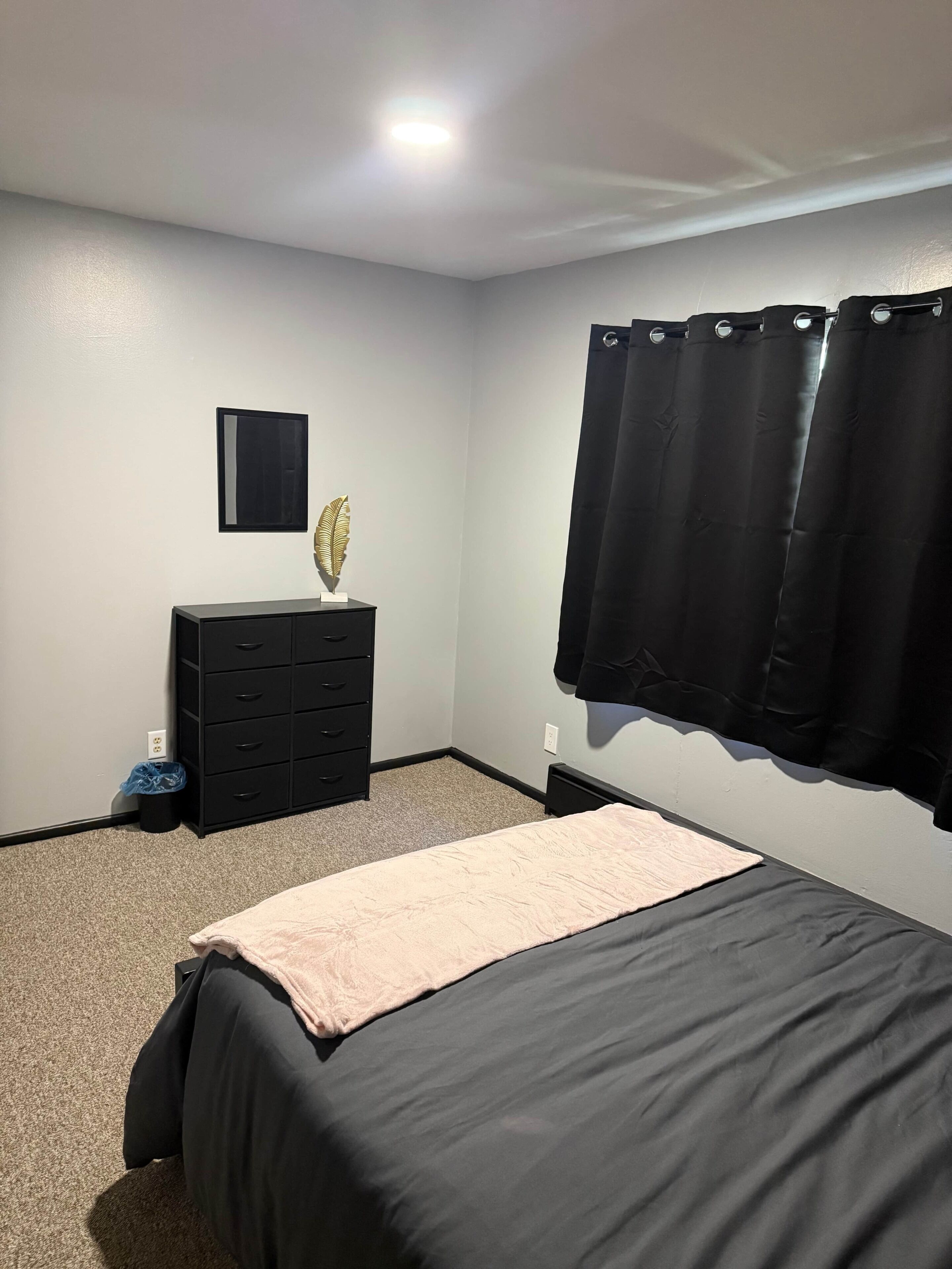 Een slaapkamer met een bed, een ladekast, een wandlamp en een raam met gordijnen.