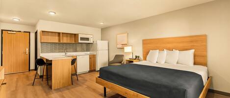 Habitación de hotel con cama de madera, una kitchenette pequeña, un escritorio con silla y una mesita de noche con lámpara.