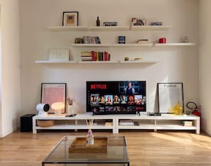 Ein Wohnzimmer mit Flachbildfernseher, weißen Regalen mit Büchern und Dekoration, einem gerahmten Bild und einem Glastisch.