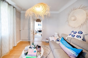 En stue med sofa, sofabord med bøger og en flaske, en hængende lampe og udsigt til et andet rum.