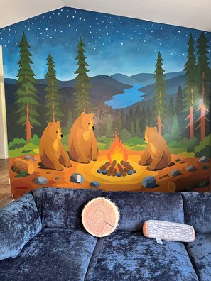 A room with a large wall mural of bears around a campfire, a blue sofa with a tree stump, and a wooden log.