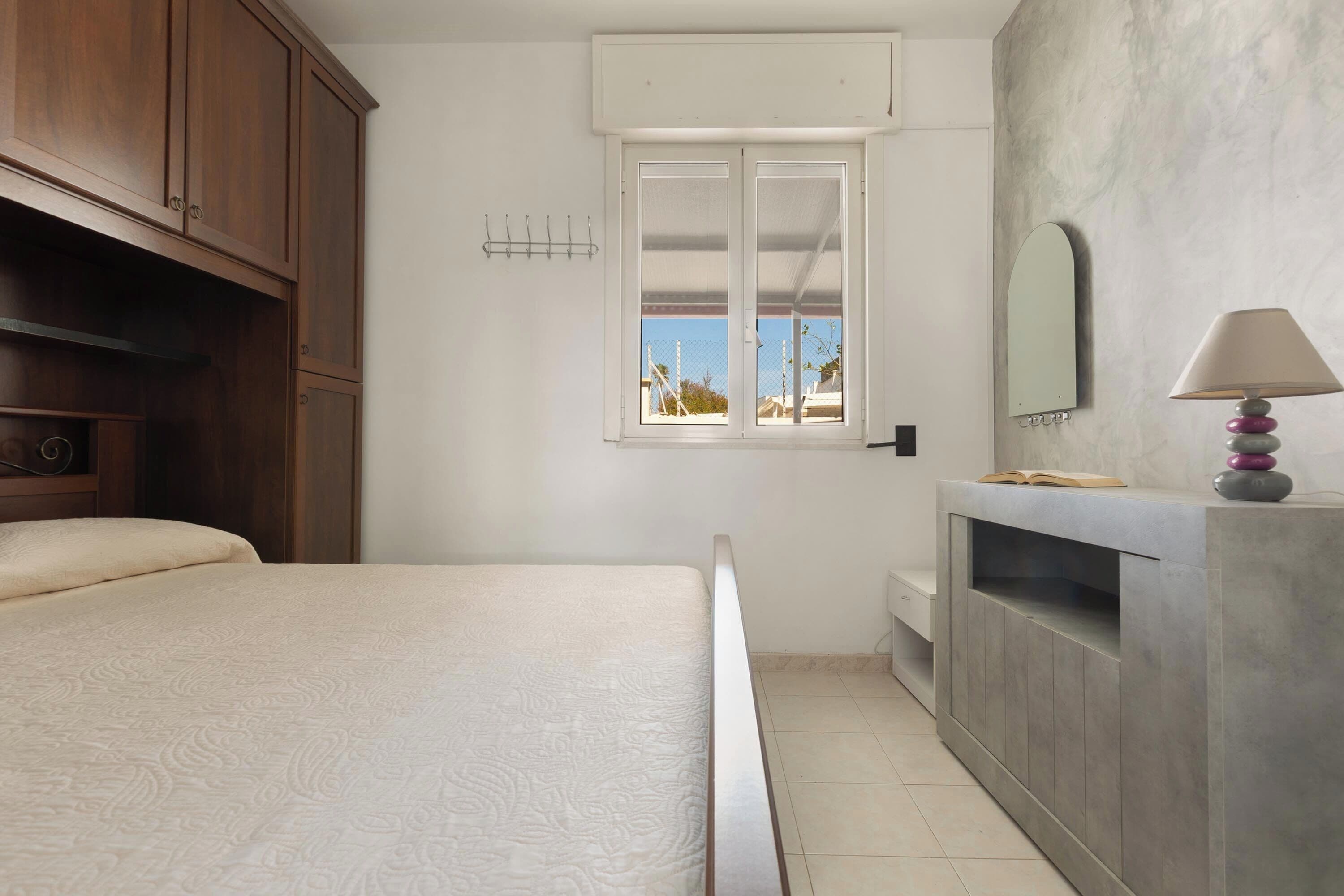 Un dormitorio con cama, ventana, espejo y mesita de noche.