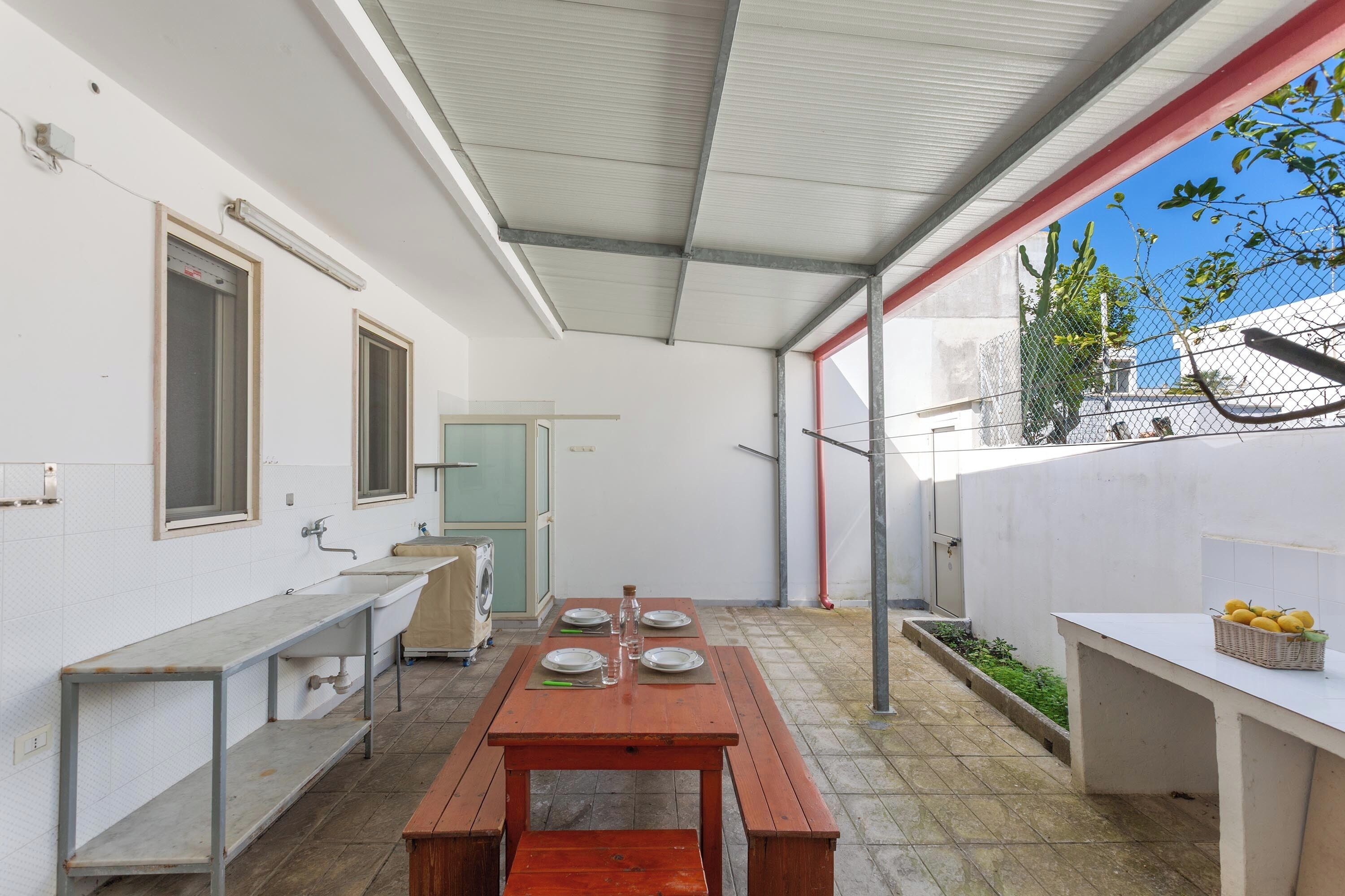 Un patio exterior cubierto con mesa de comedor, bancos y una pequeña cocina con lavamanos y lavadora.