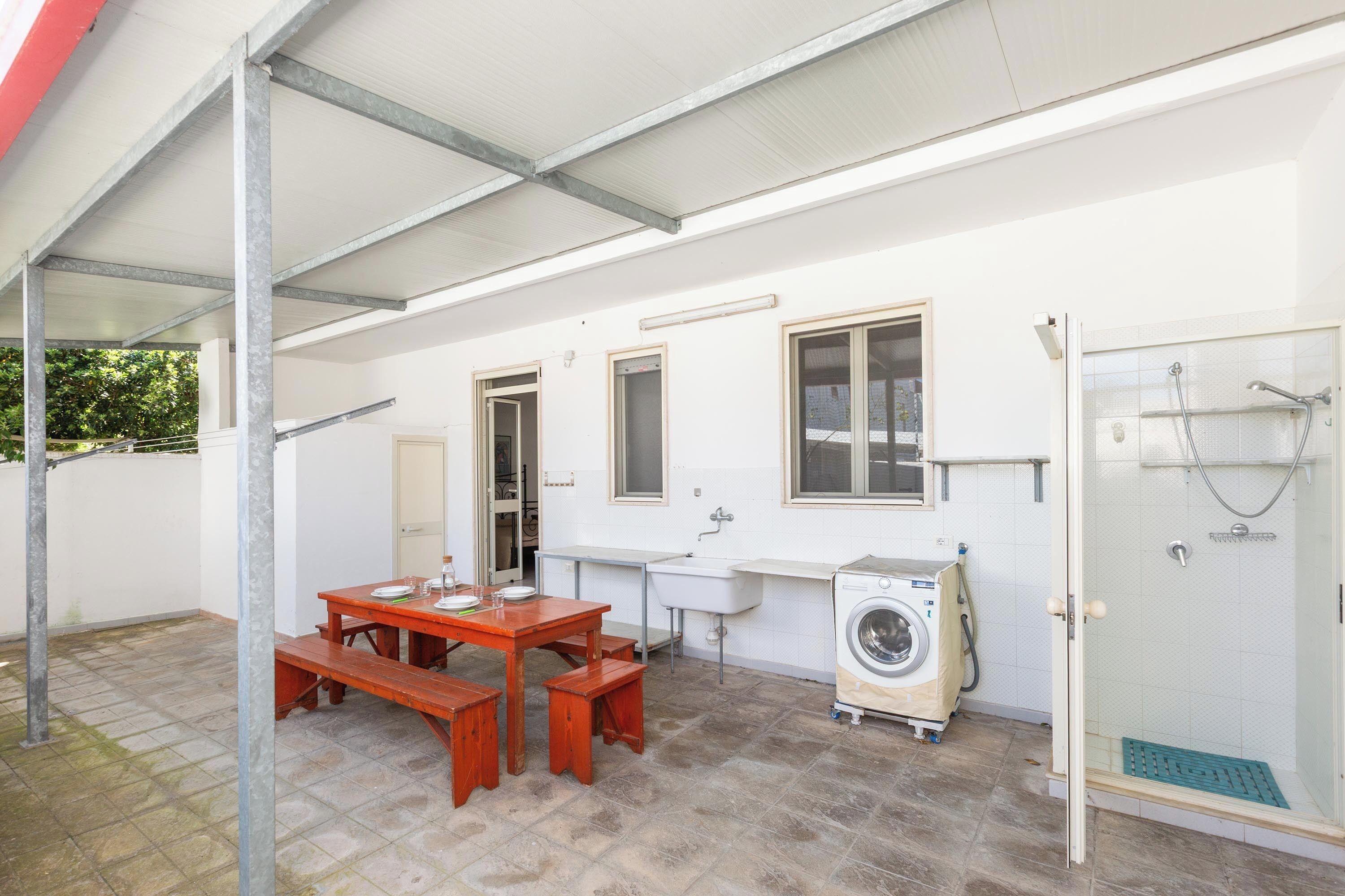 Un espace extérieur abrité comprenant une table, des bancs, un lave-linge, un évier et une douche.
