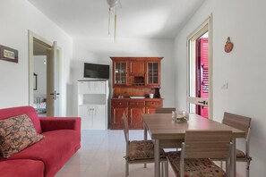 Ein Wohnzimmer mit einem roten Sofa, einem Holzessttisch mit Stühlen, einer Küchenzeile mit Holzschränken und einem Fernseher auf einem weißen Schrank.