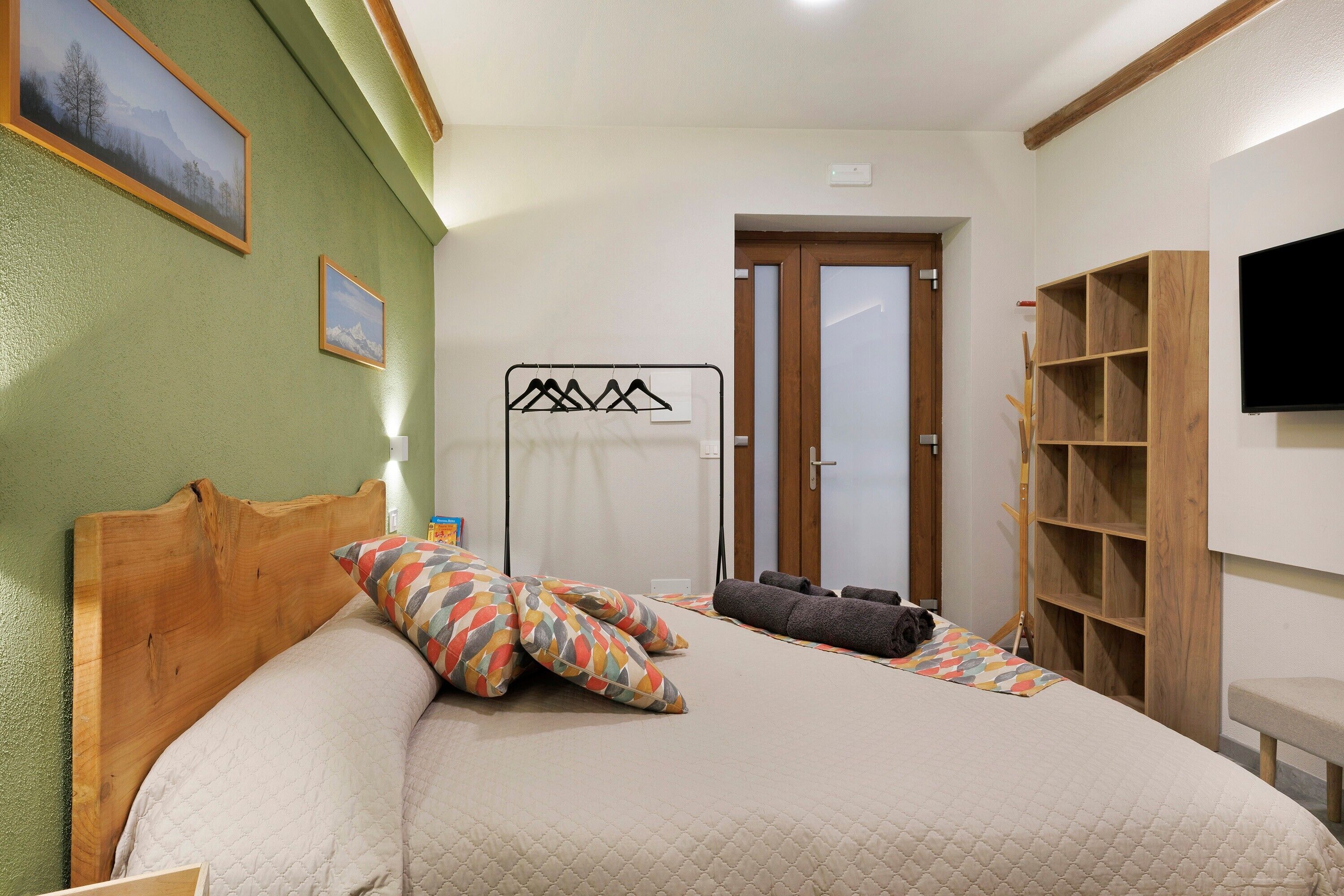 A bedroom with a wooden headboard, a bed with patterned pillows, a towel rack, a wooden wardrobe, and a television on the wall.