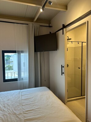 Ein Schlafzimmer mit Bett, einem Vorhangfenster, einem an der Wand befestigten Fernseher und einer Duschkabine aus Glas.
