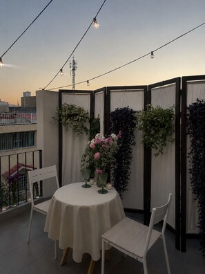 Ein Balkon mit rundem Tisch, zwei Stühlen, einer Blumenkomposition und Blick auf die Stadtlandschaft.