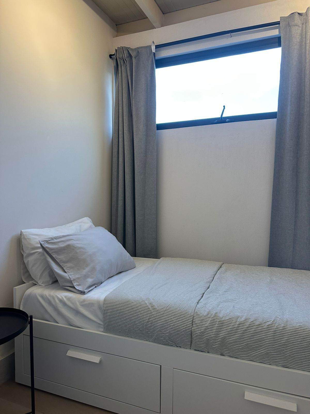 Un lit simple avec une tête de lit, une table de chevet et une fenêtre avec des rideaux.