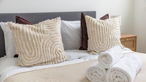 Een keurig opgemaakt bed met opgerolde witte handdoeken, twee gestructureerde kussens en een houten nachtkastje met een lamp.