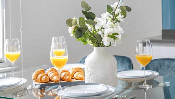 Een ontbijtsetting met croissants, sinaasappelsap en een bloemenvaas op een glazen tafel.