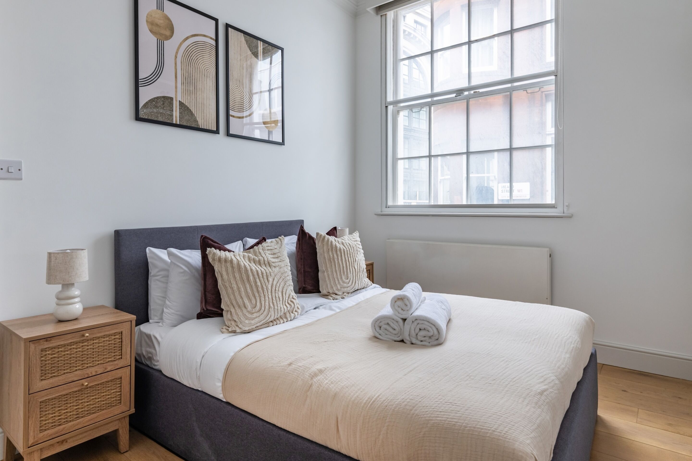 A neatly made bed with a white comforter and rolled towels, a bedside table with a lamp, and a window with a view of buildings.