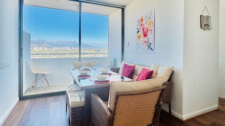 A dining area with a table set for four, a wicker chair, a sofa with cushions, and a large window with a city view.. Departamento 2001 Movistar Arena