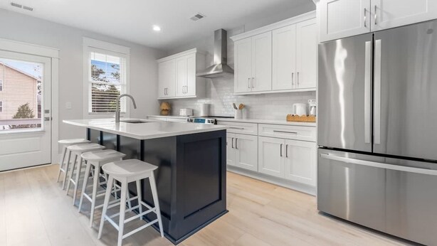 A modern kitchen with white cabinets, a stainless steel refrigerator, and a central island with stools. - The Getaway, just blocks from the ocean. (Carolina Beach)