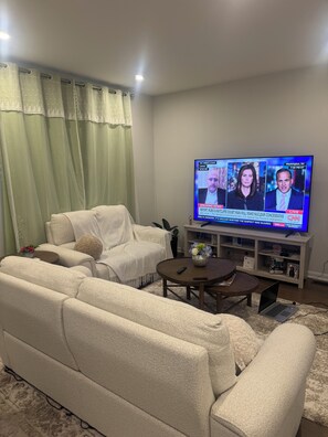 A living room with a sofa, a coffee table, a TV displaying a news broadcast, and a bookshelf. - Charming One‑Bedroom Getaway in a Peaceful Townhome Setting (Ottawa)