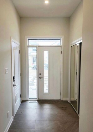 Un couloir étroit, avec une porte blanche au fond, une fenêtre au-dessus et une porte sur la gauche.