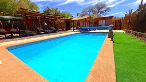 Une piscine avec un bord carrelé, une zone herbeuse et un espace de repos couvert équipé de chaises.