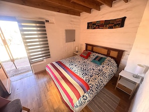 A bedroom with a bed, a bedside table, a nightstand, a window with blinds, and a wall-mounted artwork.
