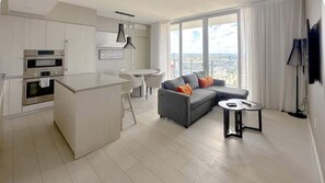 Eine moderne Küche mit integrierten Geräten, ein Essbereich mit einem weißen Tisch und Stühlen, ein graues Sofa mit orangefarbenen Kissen und ein großes Fenster mit Blick auf die Stadt.
