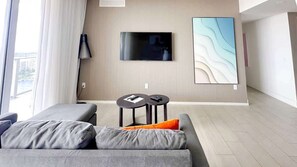 A modern living room with a grey sofa, a flat-screen TV mounted on the wall, and a small table with a lamp. - Hollywood Beach 3BR Luxe Stay Free Parking (Hollywood)