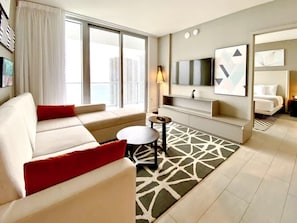 Ein modernes Hotelzimmer mit einer Couch, einem kleinen runden Tisch, einem an der Wand montierten Fernseher und einem großen Fenster mit Blick auf Gebäude.