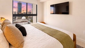 Ein Hotelzimmer mit einem Bett, einem Fernseher, einem Fenster mit Blick auf die Stadt und einer Lampe.