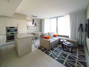 Ein modernes Hotelzimmer mit einer Küchenzeile, einem Sofa mit orangen Kissen, einem runden Couchtisch und einem gemusterten Teppich.