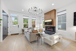 A modern living room with a fireplace, a television mounted above it, and a large window providing a view of the outside. - Linwood House | Rooftop, Hot Tub, Sleeps 15 | (Columbus)
