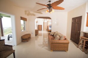 A living room with a ceiling fan, a sofa, a wicker chair, and a wooden door. - Charming 1-bedroom villa atop Bordeaux Mountain (Coral Bay)