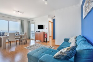 Ein modernes Wohnzimmer mit einem blauen Sofa, einem Essbereich mit Tisch und Stühlen, einem an der Wand befestigten Flachbildfernseher und Blick durch große Fenster.