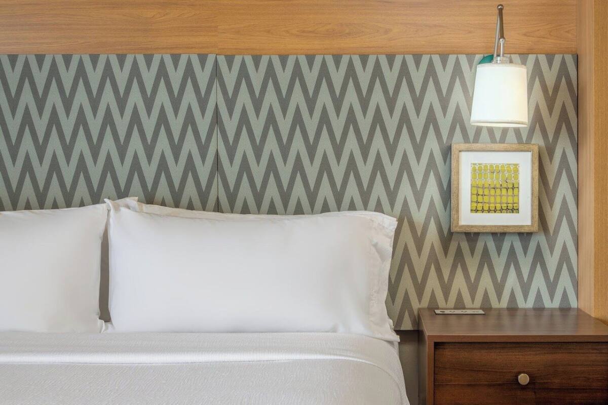 Un lit bien fait, avec du linge de lit blanc, une tête de lit en bois, une table de chevet sur laquelle est posée une photo encadrée, et un mur orné d’un motif en chevrons.