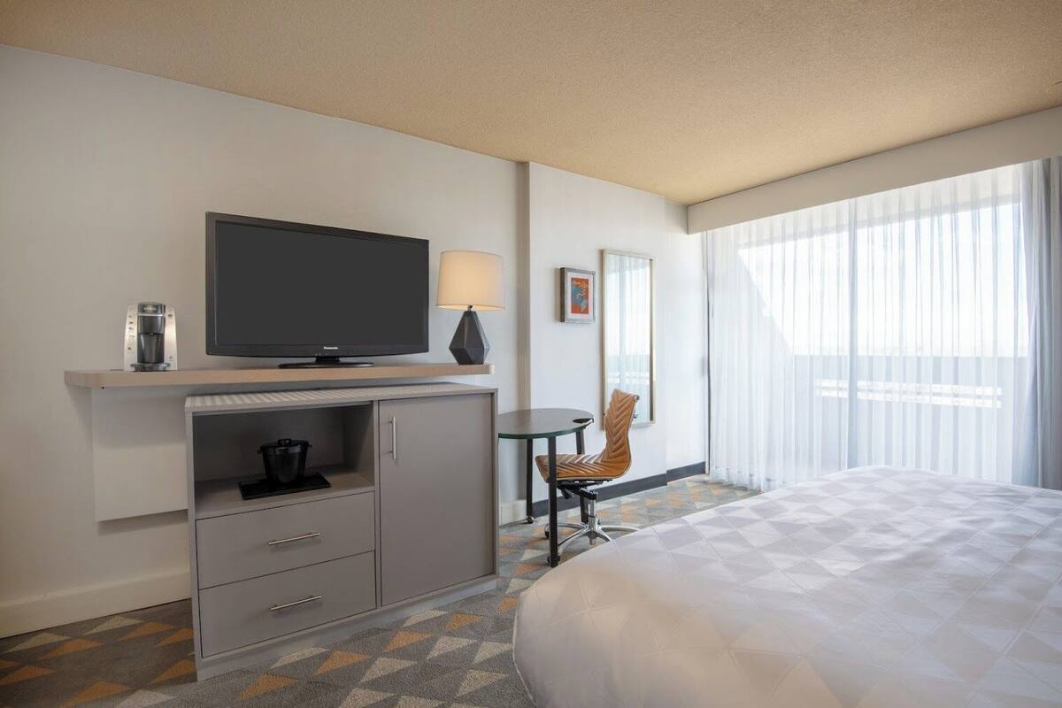Une chambre d’hôtel équipée d’un lit, d’une télévision, d’une machine à café, d’une lampe, d’une chaise et d’une fenêtre avec des rideaux.