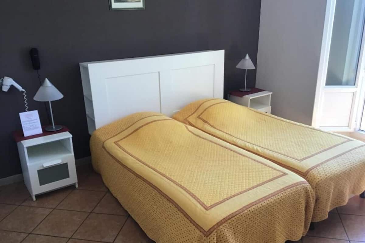 Una habitación con dos camas, una mesita de noche con lámpara, un teléfono y un estante pequeño.