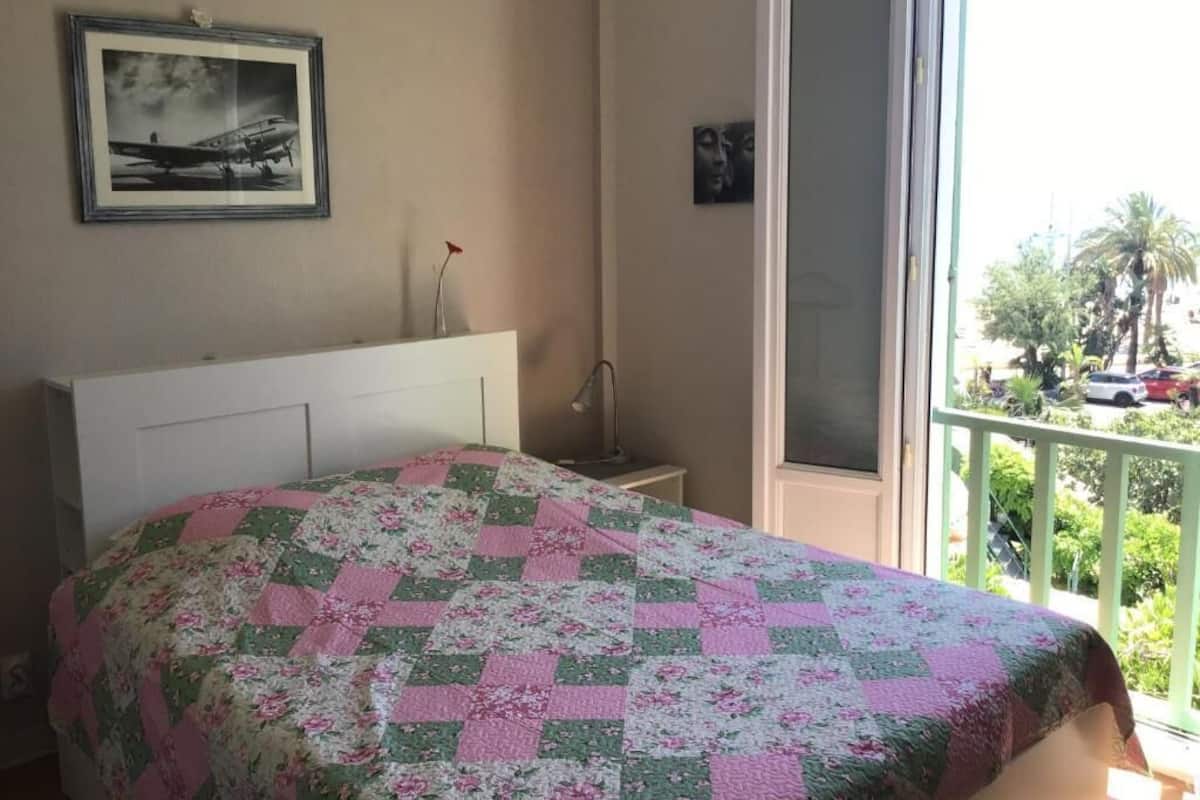 Un dormitorio con cama, un cuadro enmarcado en la pared, una mesita de noche y un balcón con vistas a palmeras y coches aparcados.