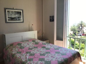 Un dormitorio con cama, un cuadro enmarcado en la pared, una mesita de noche y un balcón con vistas a palmeras y coches aparcados.